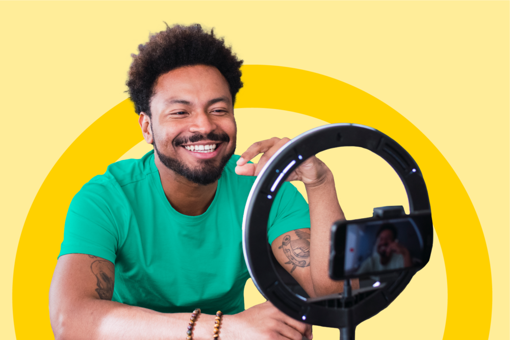 A smiling man is looking into his phone which is standing on a tripod with a ring light